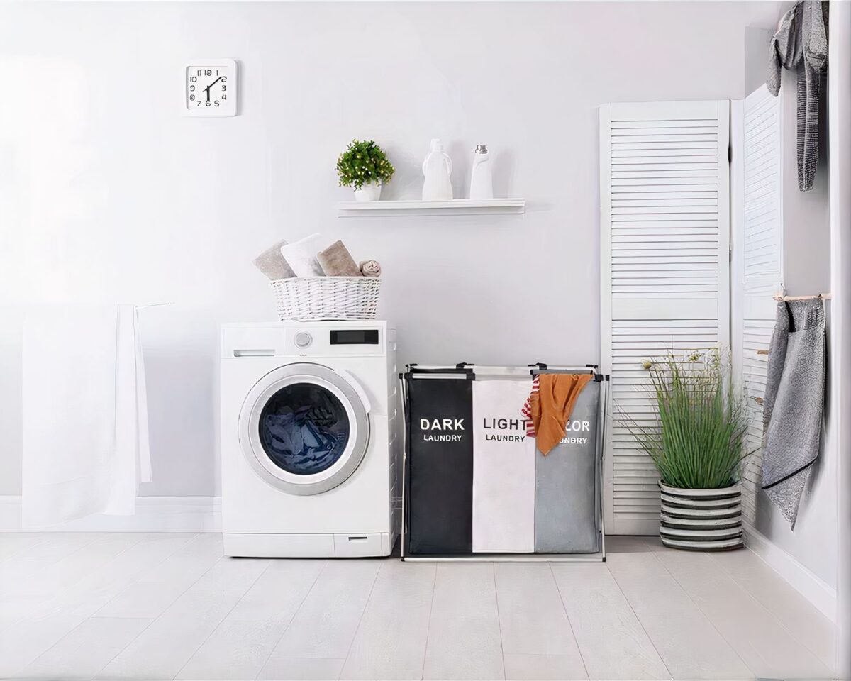 Dans une buanderie, on voit une machine à laver à côté d'une panier à linge trois compartiments en tissu.