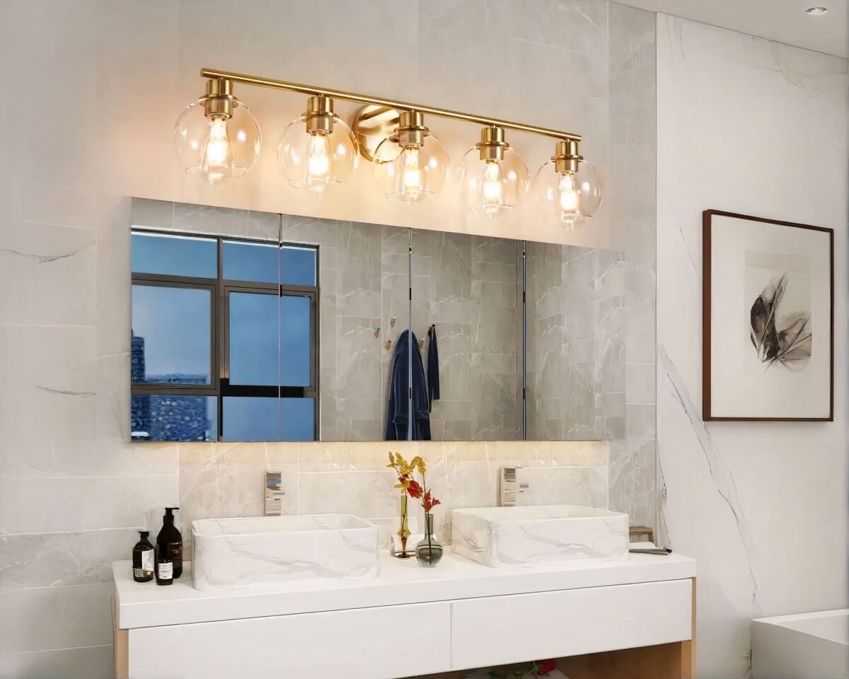 Photo représentant une applique murale de salle de bain dorée à cinq boules de verre au dessus d'un lavabo double vasque en marbre et d'un grand miroir rectangulaire montrant une fenêtre et un peignoir de bain gris dans une pièce en revêtement pierre crème