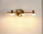 Photo d'une applique dorée pour salle de bain à trois spots rotatifs allumés sur un mur beige