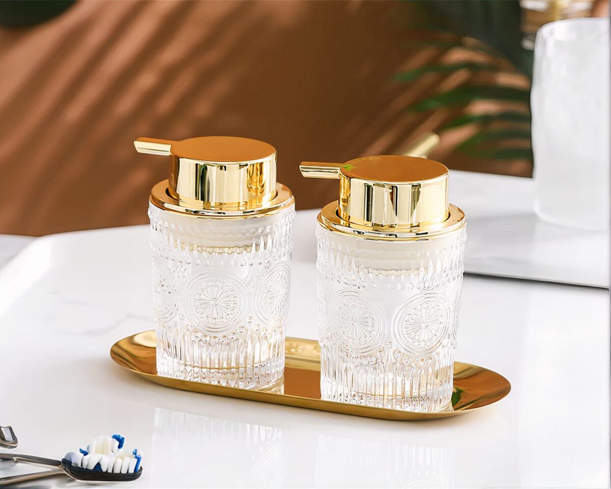 On voit deux distributeurs de savon liquide doré et verre transparent, sur un plateau.