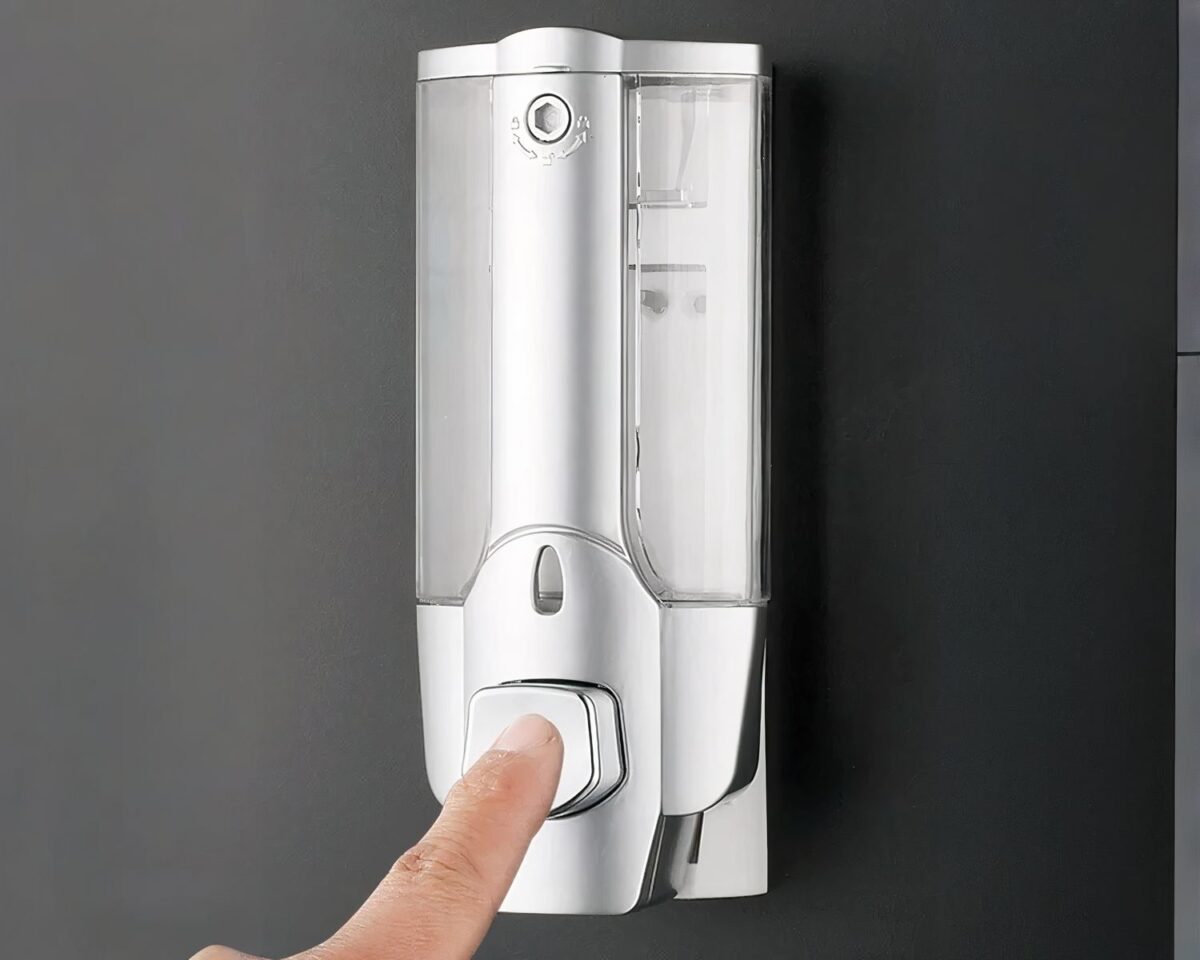On voit un doigt qui appuie sur le bouton d'une distributeur de savon mural argenté