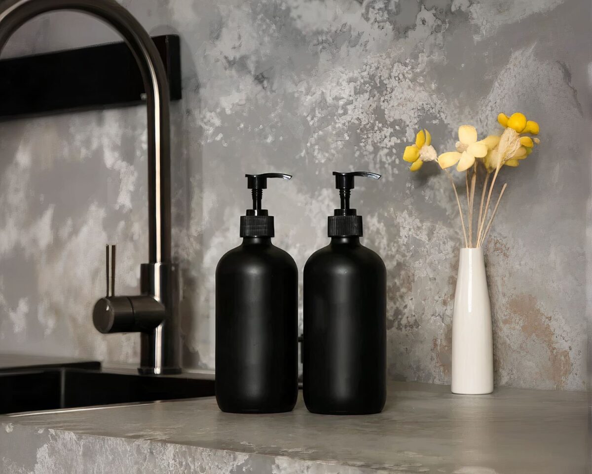 On voit deux bouteilles noires mates, des distributeurs de savon, près d'un évier dont la crédence semble en marbre. Fleurs jaunes