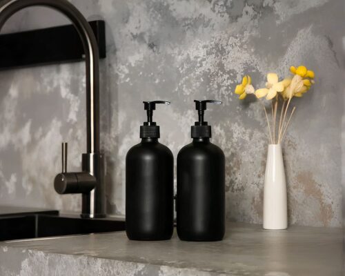 On voit deux bouteilles noires mates, des distributeurs de savon, près d'un évier dont la crédence semble en marbre. Fleurs jaunes