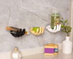Une patère blanche, une dorée, une noire en forme de main collées au mur avec des accessoires de salle de bain dans les mains