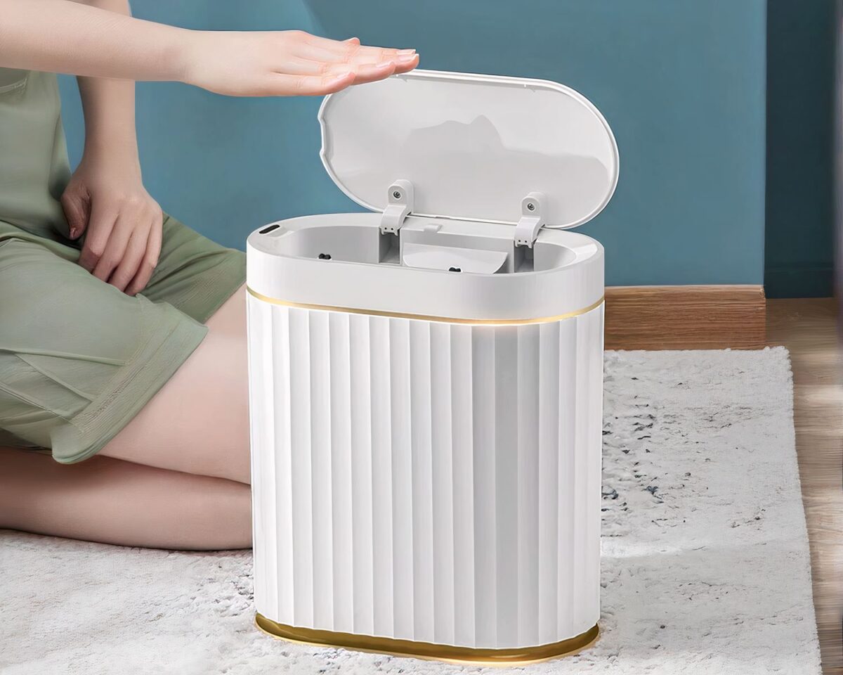 Photo d'une poubelle blanche au design ondulé à liserés dorés ouverte avec une main de femme sur le couvercle, jeune femme assise sur le sol genoux pliés et l'autre main sur sa jupe verte, le sol est clair style marbré avec une plinthe marron au fond et mur vert