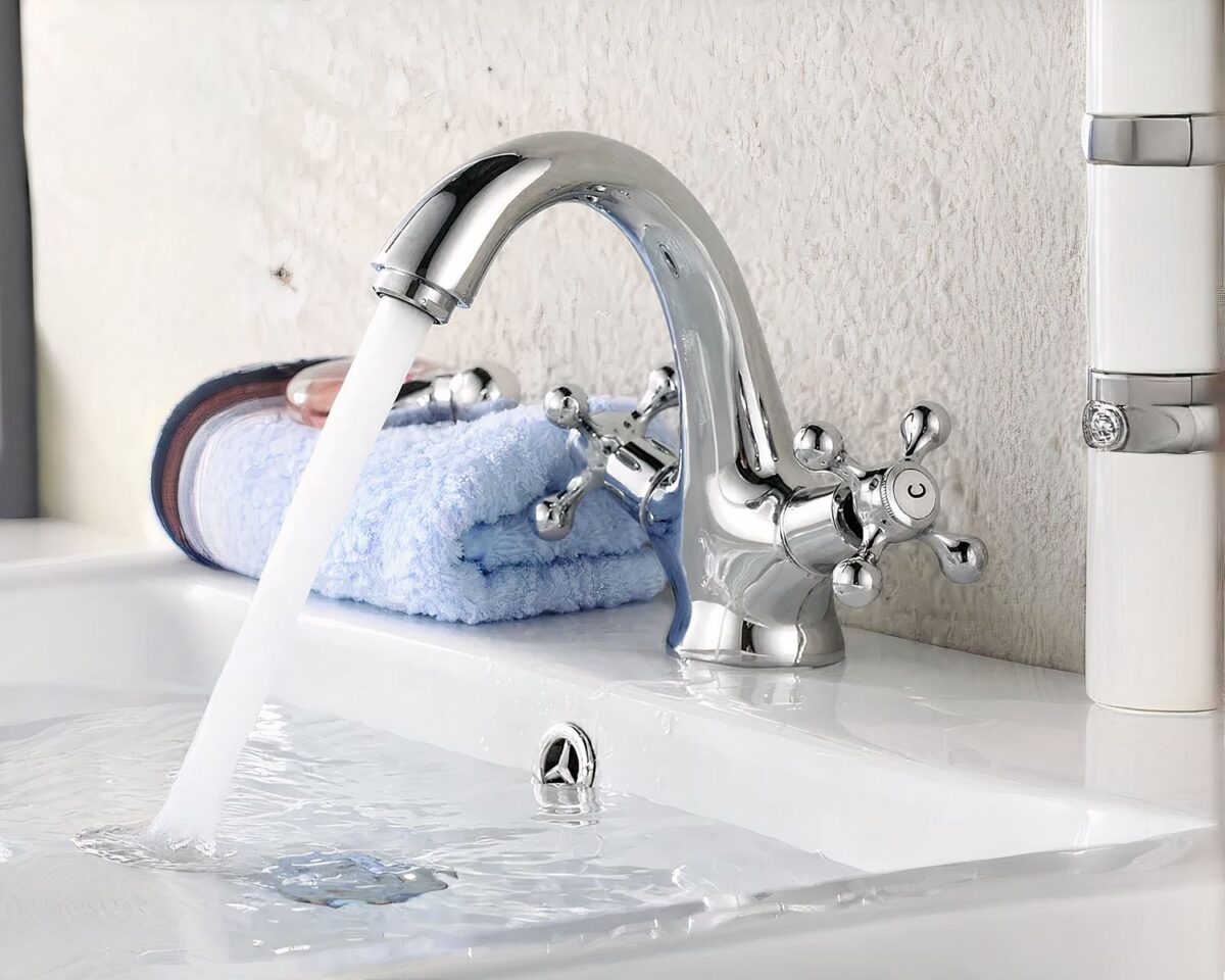 Photo d'un robinet de salle de bain argenté à deux poignées ouvert monté sur un lavabo blanc rempli d 'eau avec sur la gauche une serviette pliée bleu pale le mur derrière est blanc cassé