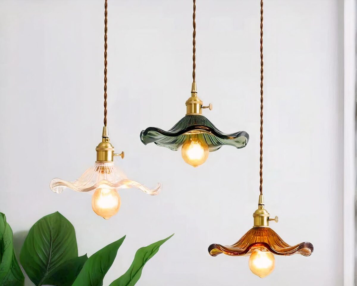 Photo représentant trois suspensions de salle de bain une transparente, une verte et une marron en verre allumées avec de grosses ampoules et de longueur différente devant un mur gris pale et un haut de plante verte sur la gauche