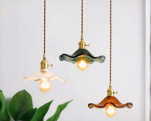 Photo représentant trois suspensions de salle de bain une transparente, une verte et une marron en verre allumées avec de grosses ampoules et de longueur différente devant un mur gris pale et un haut de plante verte sur la gauche